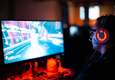 A person wearing headphones is sitting in front of a computer monitor. The screen displays a vibrant, illuminated video game scene, while the person appears focused on gaming. The ambient lighting is dark with red and blue tones, and a backlight from the monitor highlights the headphones.