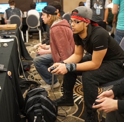 Several people are sitting in a room with gaming consoles and monitors, focusing intently on their screens. The setting appears to be a casual gaming event, with participants holding controllers while playing video games. The room has a casual atmosphere with chairs and tables set up for gaming.