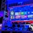 A modern gaming setup featuring a large wall-mounted screen displaying a gaming console interface. The room is decorated with gaming-themed items such as controllers and collectibles. The ambient lighting includes shades of blue and red, creating a vibrant atmosphere.